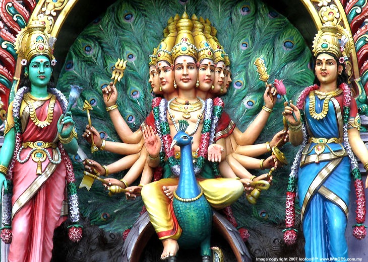 Large statue of Lord Murugan (also known as Kartikeya or Skanda) at Batu Caves in Malaysia, a major deity in Tamil Hindu tradition associated with youth, wisdom, and the defeat of ignorance. Murugan is shown with multiple arms holding symbolic weapons and emblems, signifying diverse powers and forms of knowledge. He stands with or near his peacock mount, which traditionally represents the conquest of destructive impulses and ignorance; serpent imagery in Murugan iconography is often read as the subjugation of ignorance and ego. The statue is richly ornamented and monumental, set within the Batu Caves pilgrimage complex.